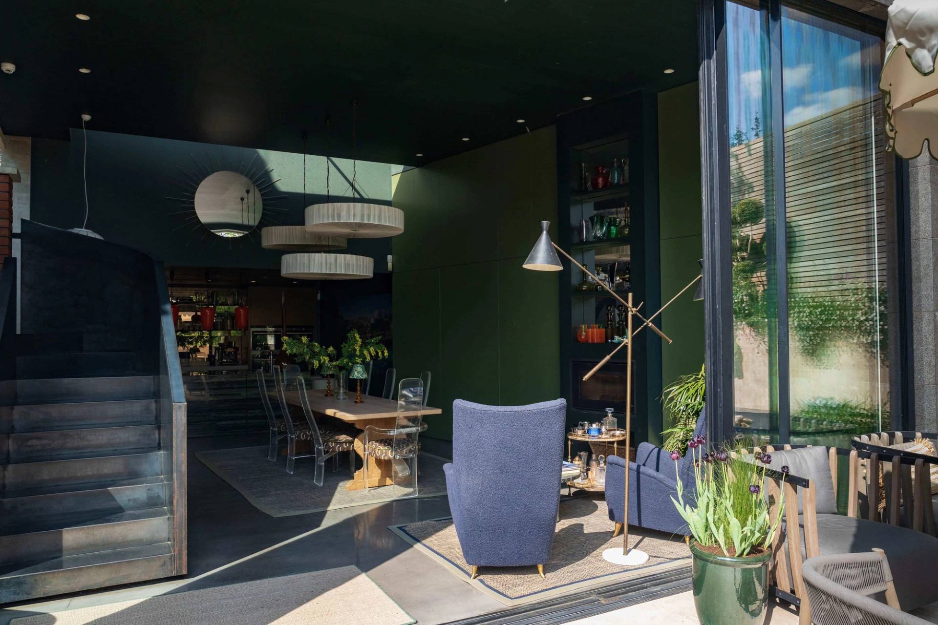 Sunlit architectural lounge zone with floor-to-ceiling glass walls, blue armchairs, and views onto a paved garden terrace in Wimbledon, Love Locations SW20.