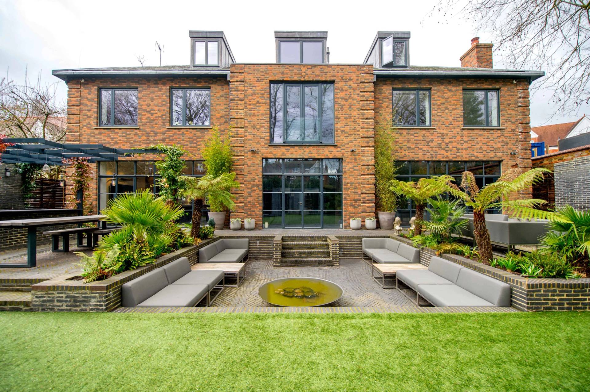 Landscaped rear patio terrace featuring sunken outdoor sofa seating around a central fire pit, Love Locations.