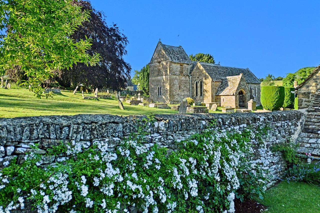 Historic Cotswold stone church and graveyard shoot location in Gloucestershire GL7 from Love Locations, featuring white spring flowers in the foreground, traditional stone walls, and clear blue skies.