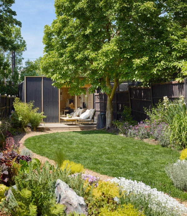 Curved grass lawn path leading through lush flower borders to a dark timber garden studio room, Love Locations E17.