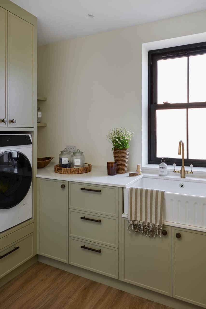Country-style utility laundry room location featuring sage green cabinetry, a white Belfast sink, and integrated appliances, ideal for home appliance commercials, Love Locations.