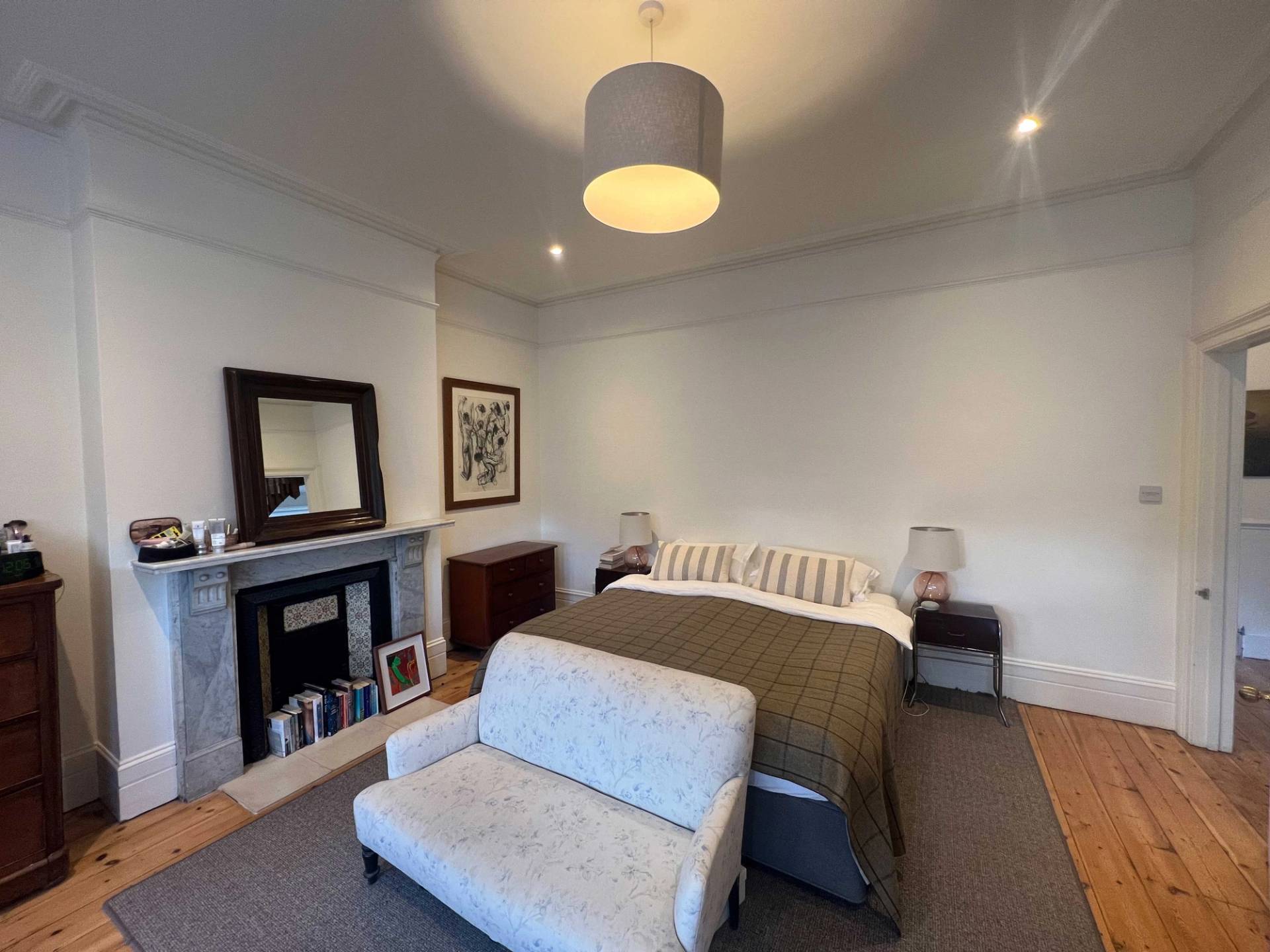 Spacious master bedroom with neutral bedding, grey upholstered bench, period fireplace, and warm carpet, Love Locations Denmark Hill.