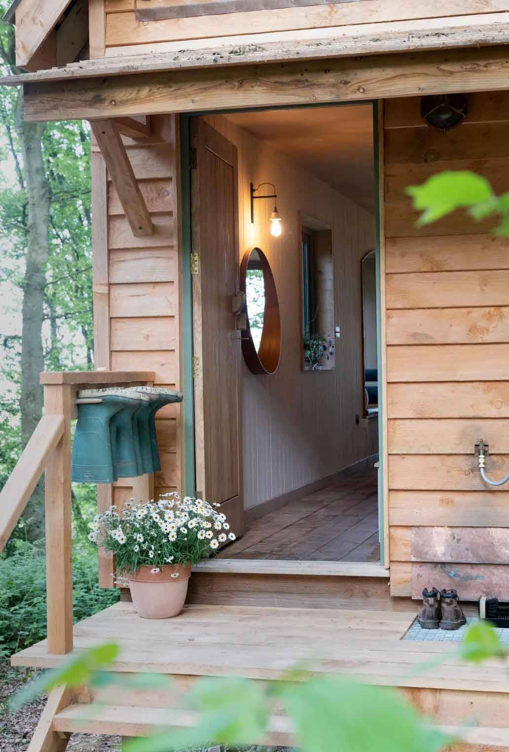 Open wooden entrance door to a timber frame treehouse showing a warm interior corridor and potted white flowers on the deck, Love Locations.