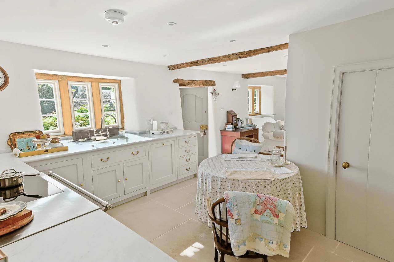 Country cottage kitchen and dining nook shoot location in Gloucestershire GL7 through Love Locations, featuring shaker-style cabinetry, exposed ceiling beams, and a round dining table.