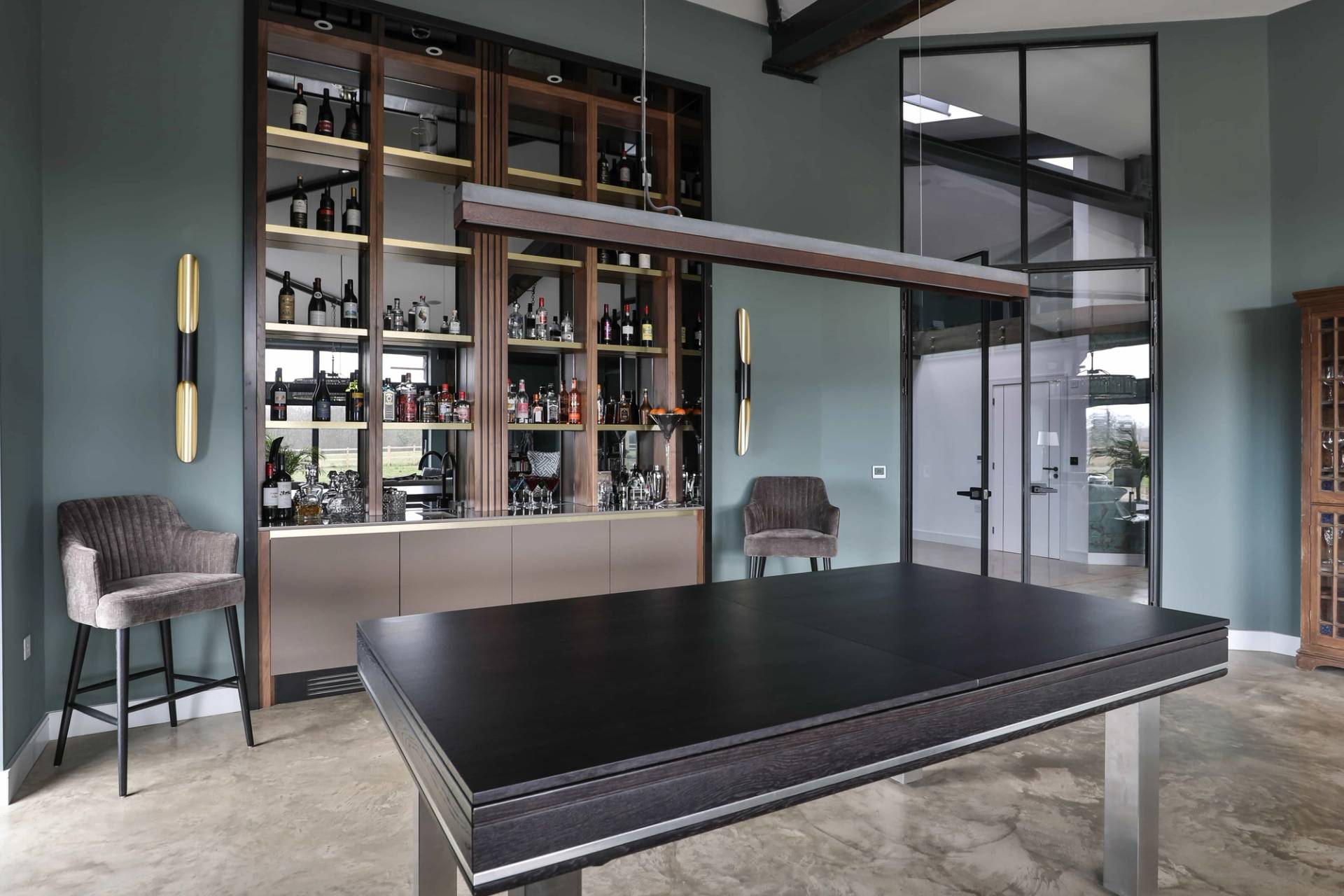 Modern bar lounge area featuring a sleek dark table, brass wall sconces, and glass bottle display shelving, Love Locations CV23.