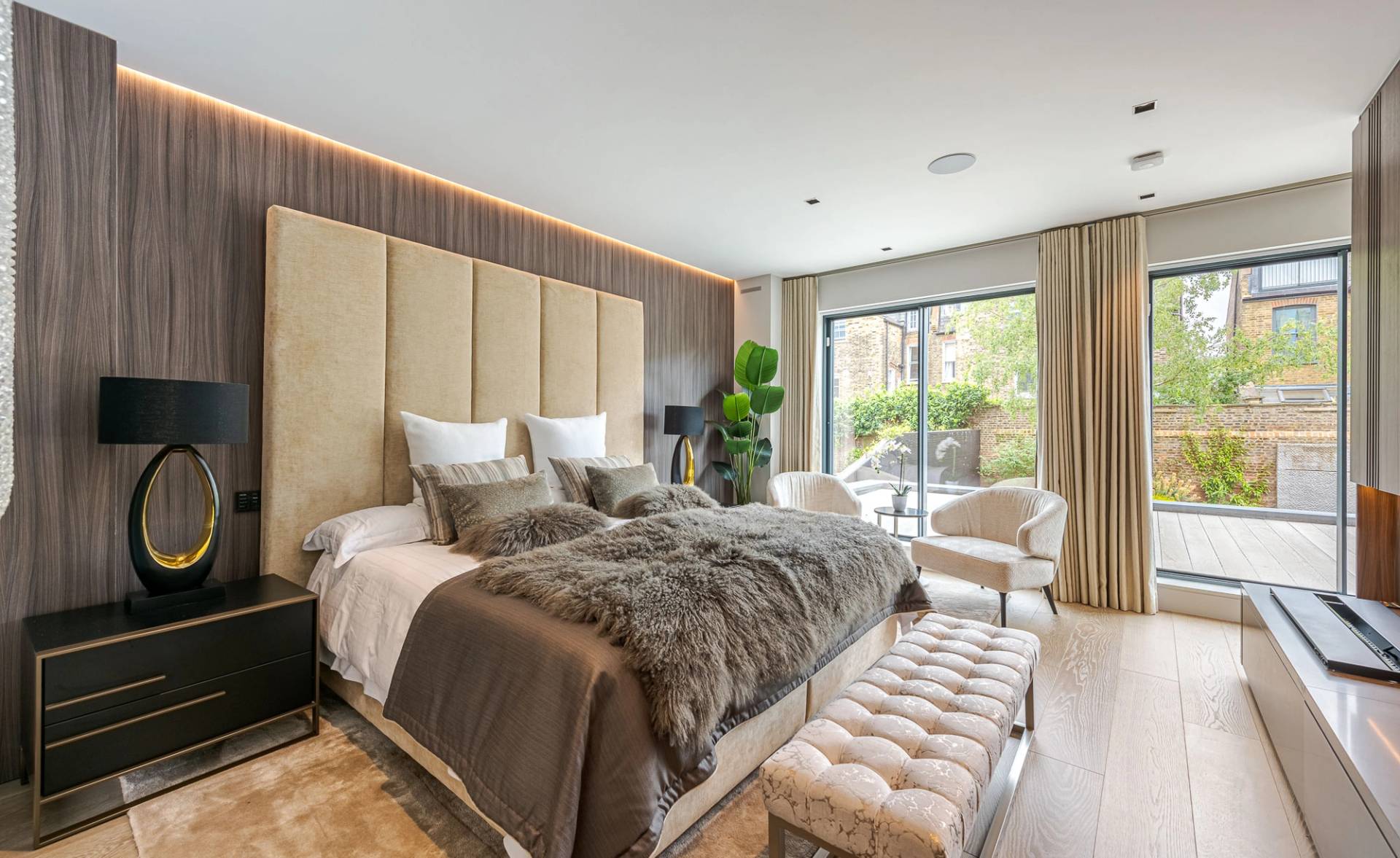 Master bedroom suite with a upholstered headboard wall, brown faux fur throw, footstool, and patio garden access, Love Locations SW11.