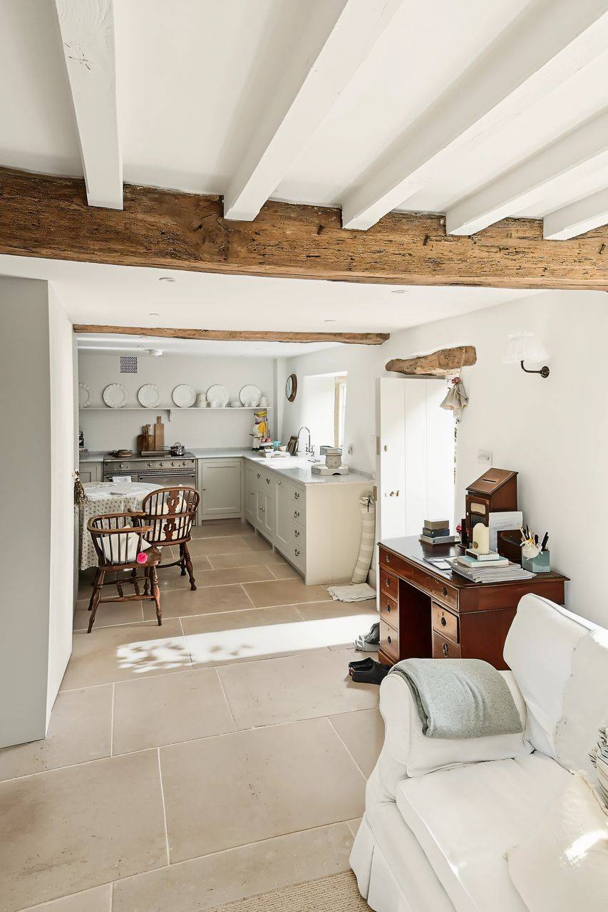 Open-plan cottage living and dining room location in Gloucestershire GL7 by Love Locations, displaying low beamed ceilings, neutral decor, and a wooden writing desk.
