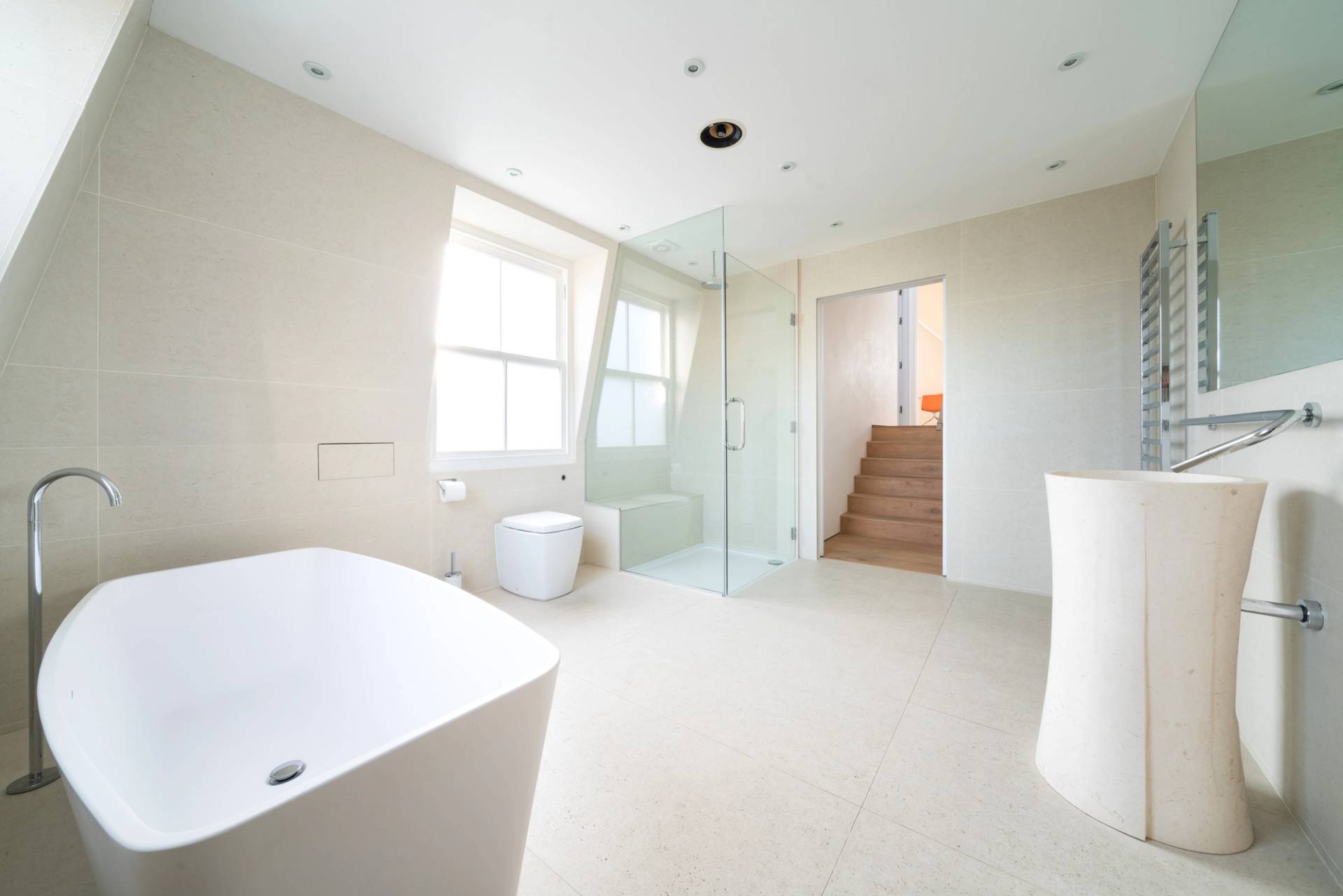 Sunlit bathroom view looking towards a freestanding white bathtub and wooden stairs, Love Locations SW6.