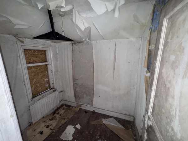 Corner of a derelict bedroom showing heavily peeling plaster sheets hanging from the ceiling near a boarded window, Love Locations.