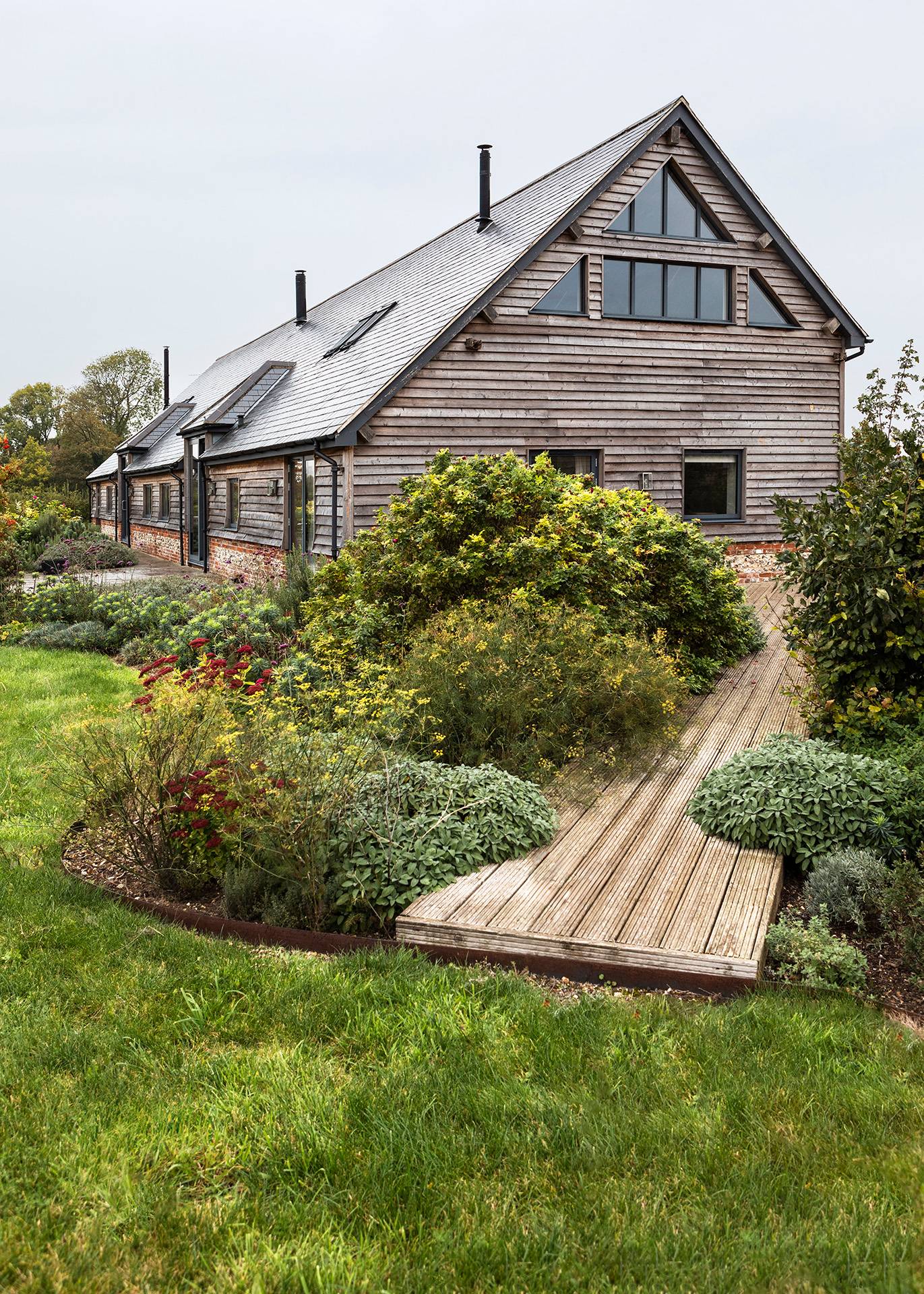 Curved timber boardwalk garden path leading through lush green lawns toward a converted barn home, Love Locations SN8.