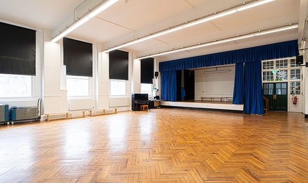 Historic school assembly hall featuring parquet wood flooring and a performance stage with blue curtains, listed on Love Locations.