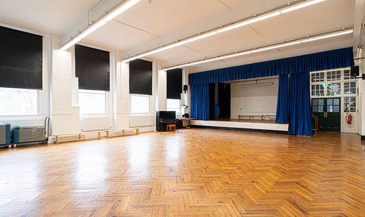 Historic school assembly hall featuring parquet wood flooring and a performance stage with blue curtains, listed on Love Locations.