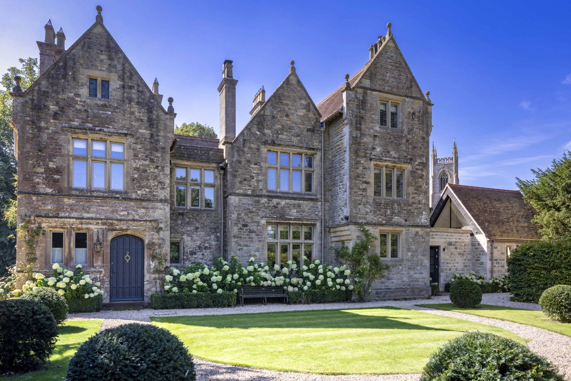 Historic Cotswold stone manor house front facade with gabled roofs and manicured lawn gardens, Love Locations OX7.