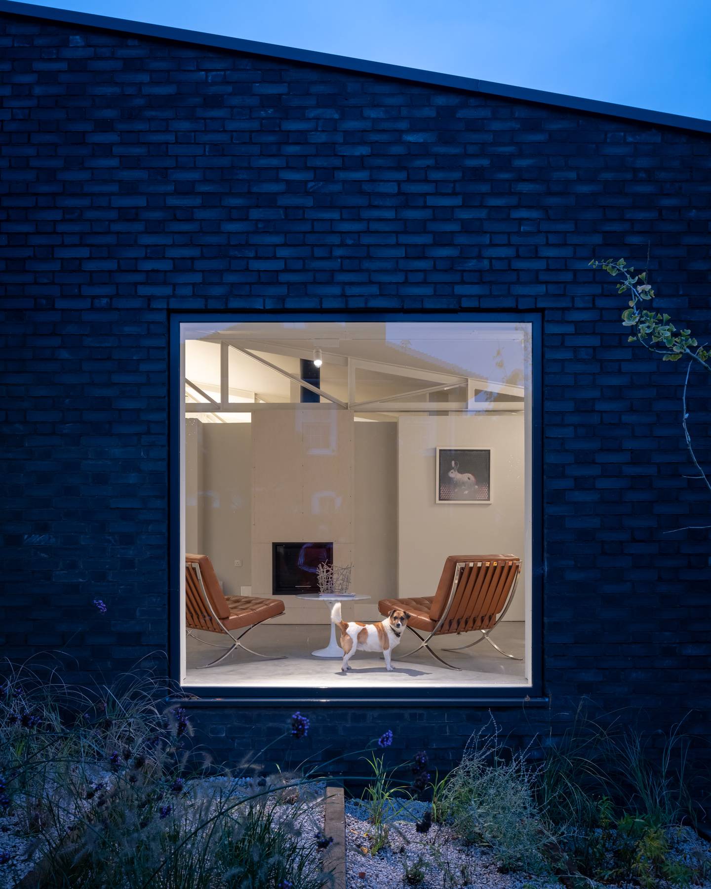 Square picture window framing cognac leather Barcelona armchairs illuminated inside a dark brick facade at dusk, Love Locations BN3.