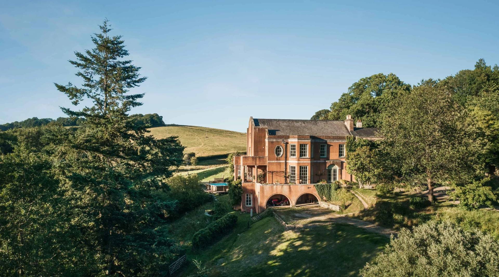 Dramatic exterior shot of a three-story red brick estate with arched porticos and lush grounds in Ipplepen Totnes Devon TQ12 from Love Locations.