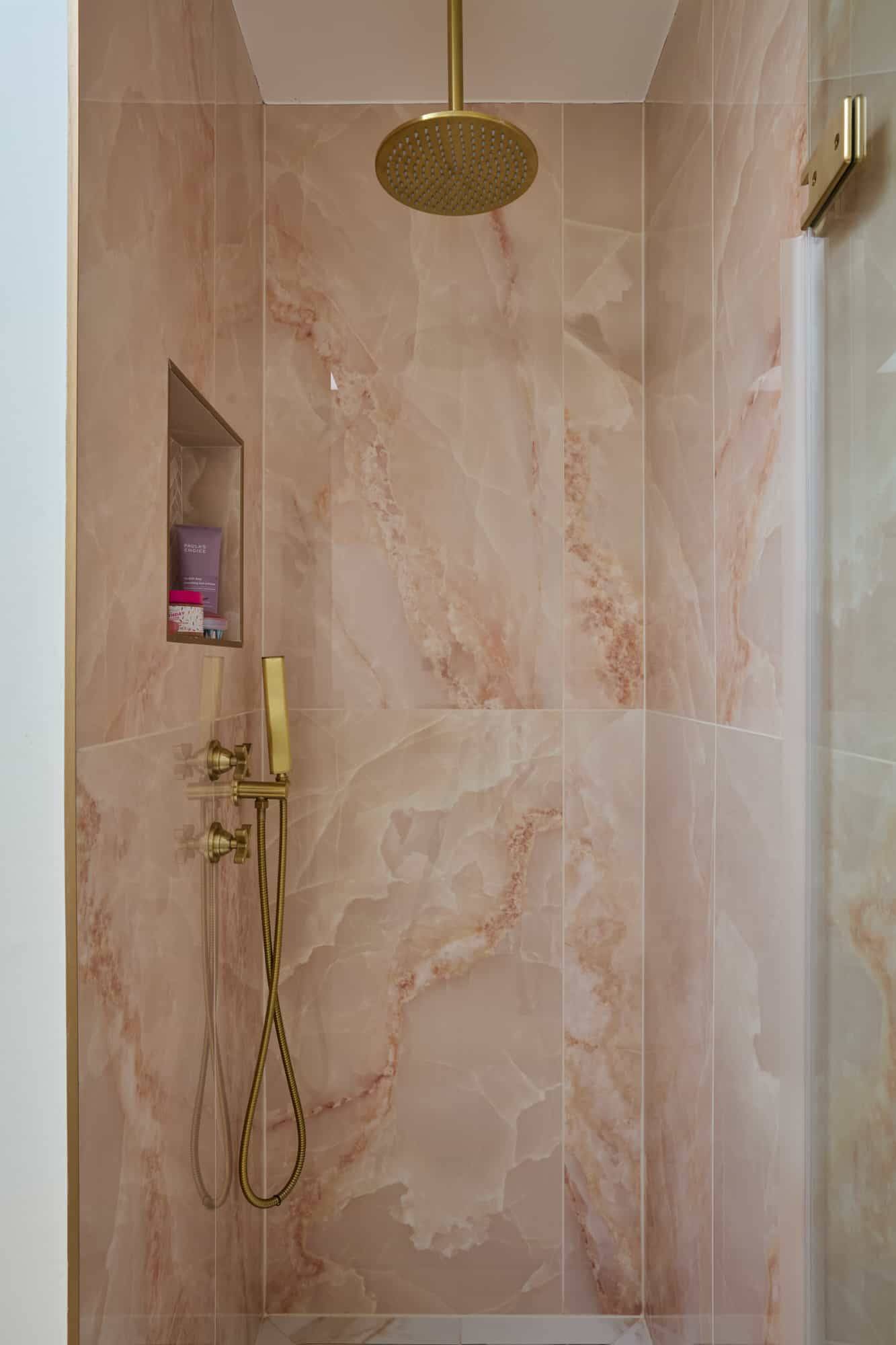 Walk-in shower enclosure lined with pink marble slab tiles and brass shower head fixtures, Love Locations.