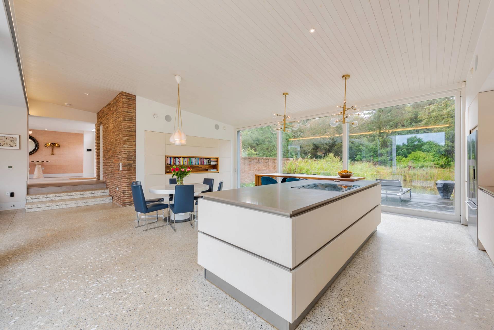 Love Locations sleek open plan kitchen GU8: Minimalist white island unit and polished terrazzo stone floors facing floor-to-ceiling glass panel windows, perfect for luxury appliance commercials.