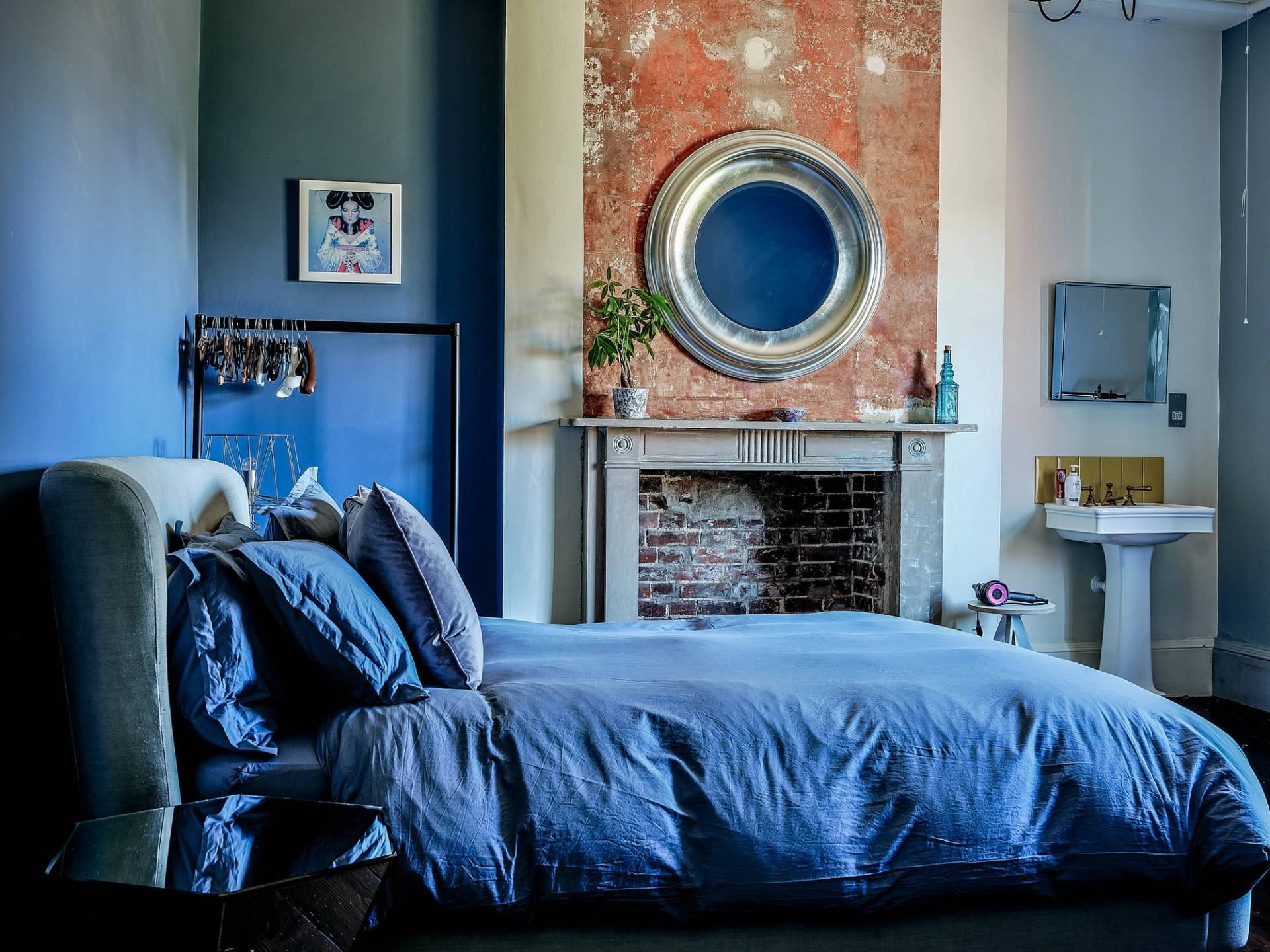 Master bedroom with exposed terracotta brick wall accents, marble fireplace, and deep blue bed linens, Love Locations.