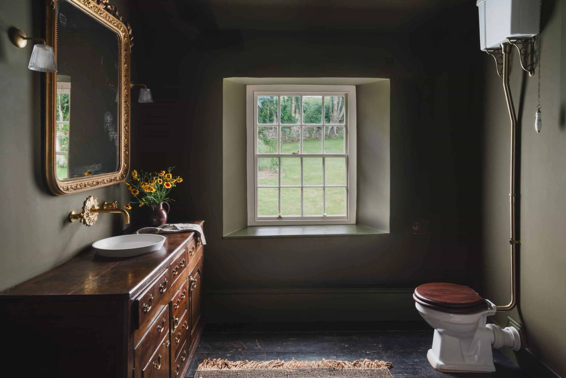 Dark moody green powder room with a wooden vanity and sash window in a historic Cornish manor by Love Locations.