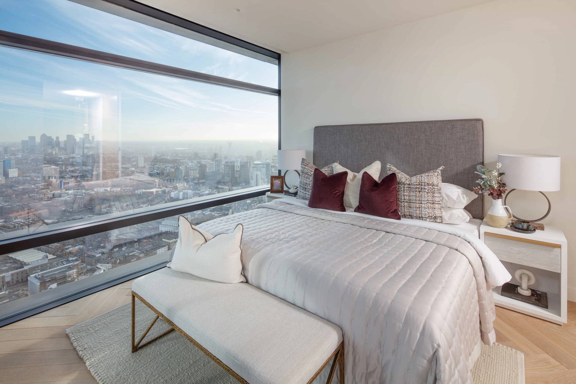 Spacious master bedroom location featuring a plush upholstered bed, white bench, and floor-to-ceiling glass wall skyline views, ideal for mattress brand advertising via Love Locations.