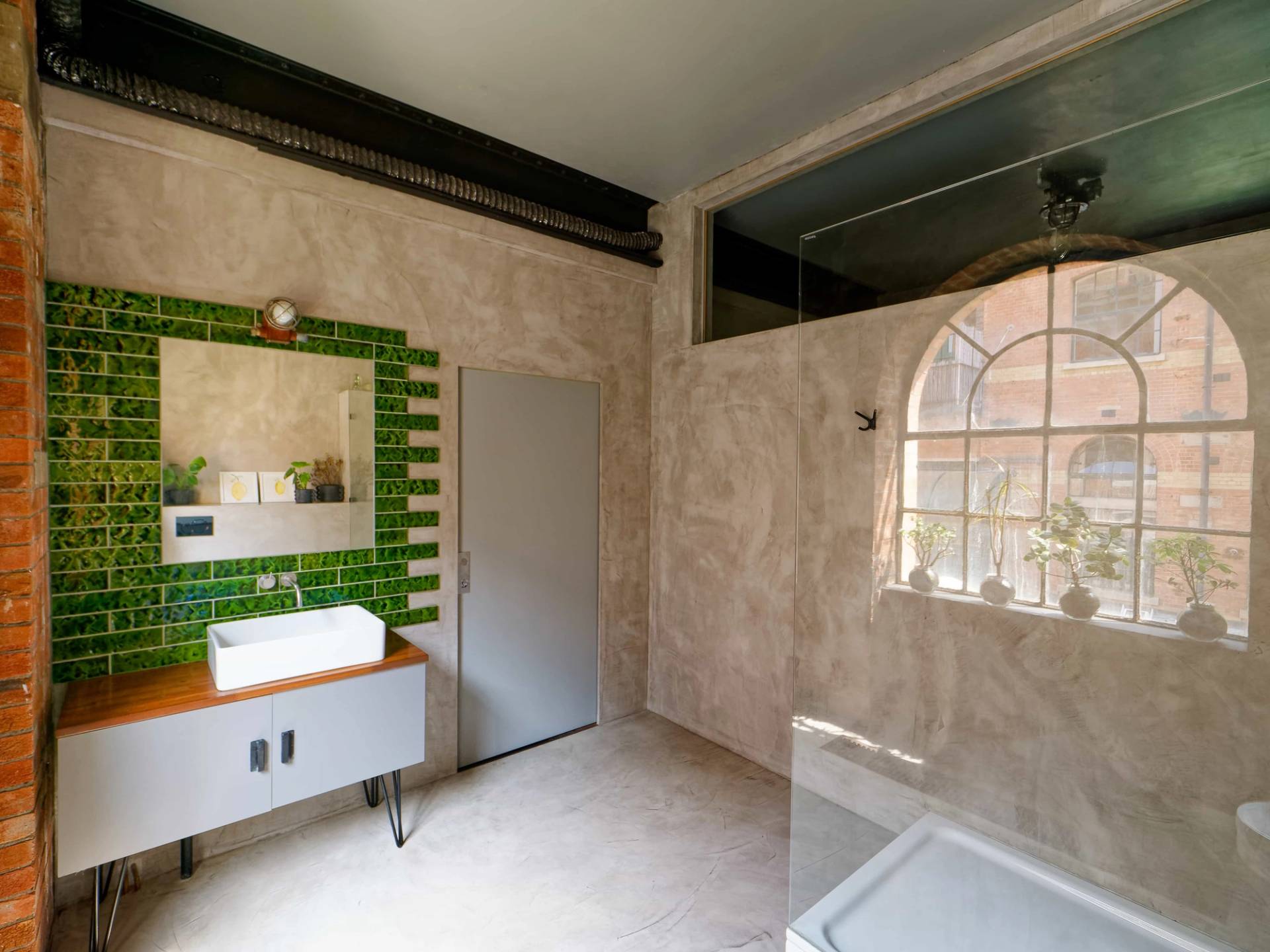 Micro-cement bathroom view showing a green tiled mirror frame, white washbasin, and walk-in shower, Love Locations E14.