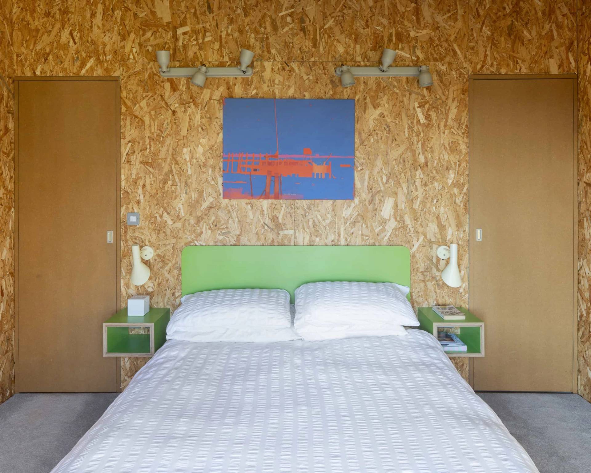 Minimalist bedroom with OSB plywood walls, lime green upholstered headboard, and bold blue canvas wall art, Love Locations IP17.