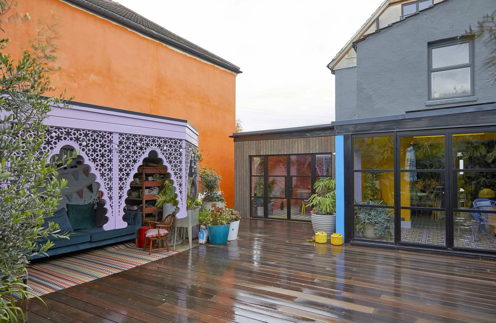 Architectural view of an outdoor timber deck flanked by orange and blue painted house exteriors and purple arches, Love Locations Catford.