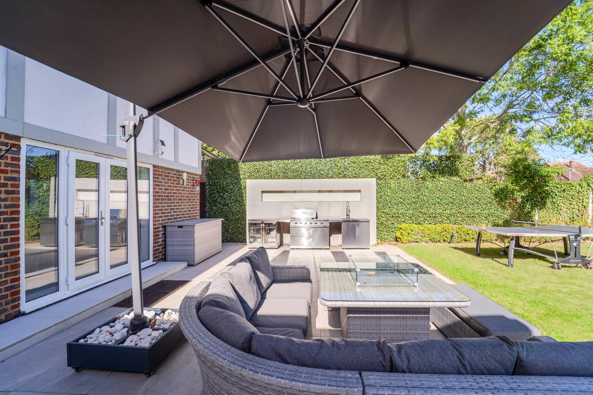 Outdoor lounge seating area set under a large black parasol beside an outdoor kitchen BBQ, Love Locations RM11.