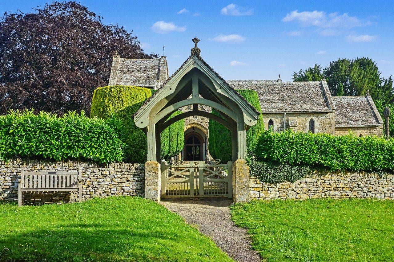 Traditional covered lychgate and stone wall entrance location house in Gloucestershire GL7 through Love Locations, with manicured hedges and a timber-framed gate structure.
