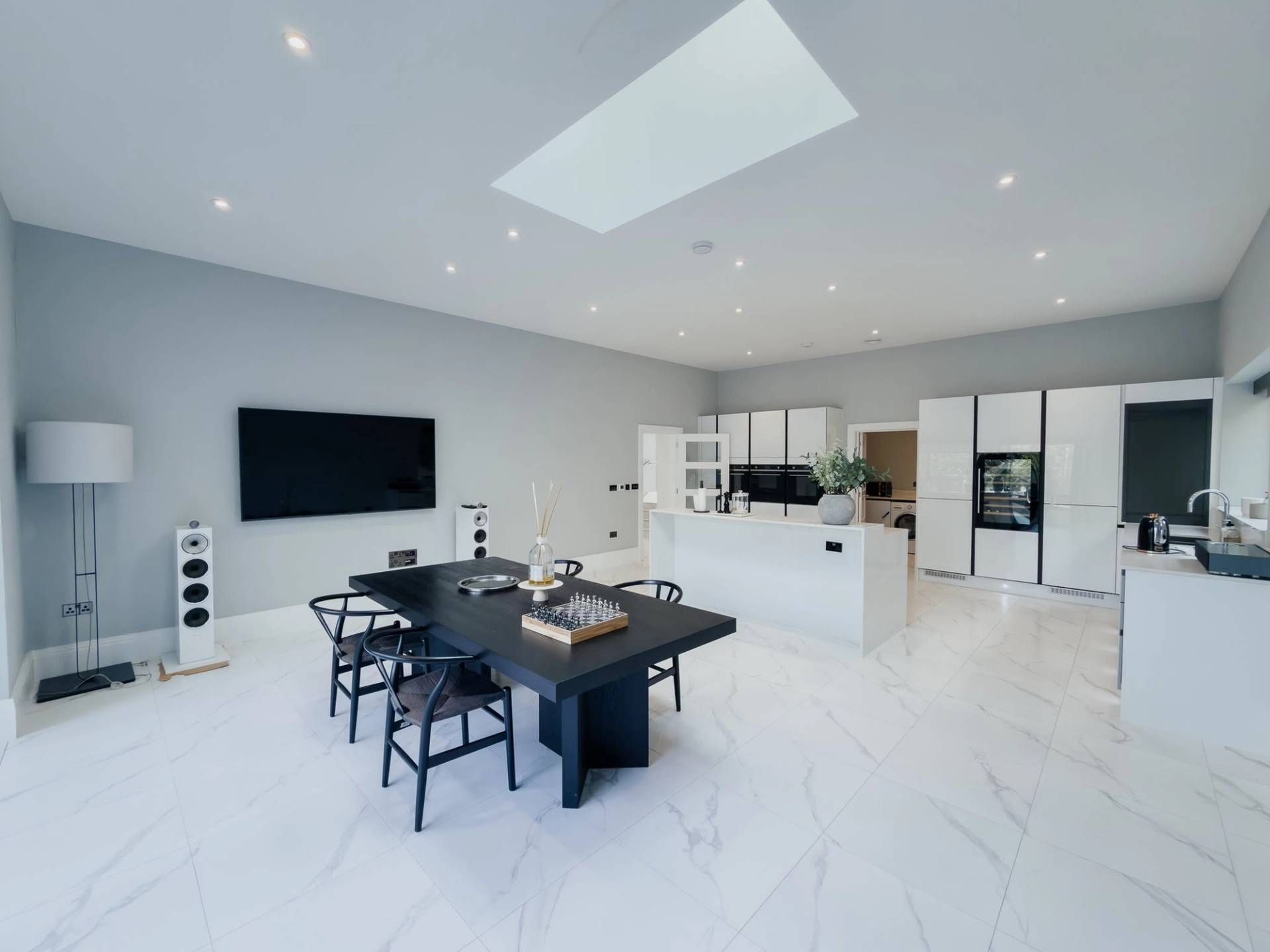 Sunlit open-plan luxury kitchen and dining room location house in Barnet EN4 from Love Locations, featuring white marble tile floors, dark dining table set, and a large skylight.