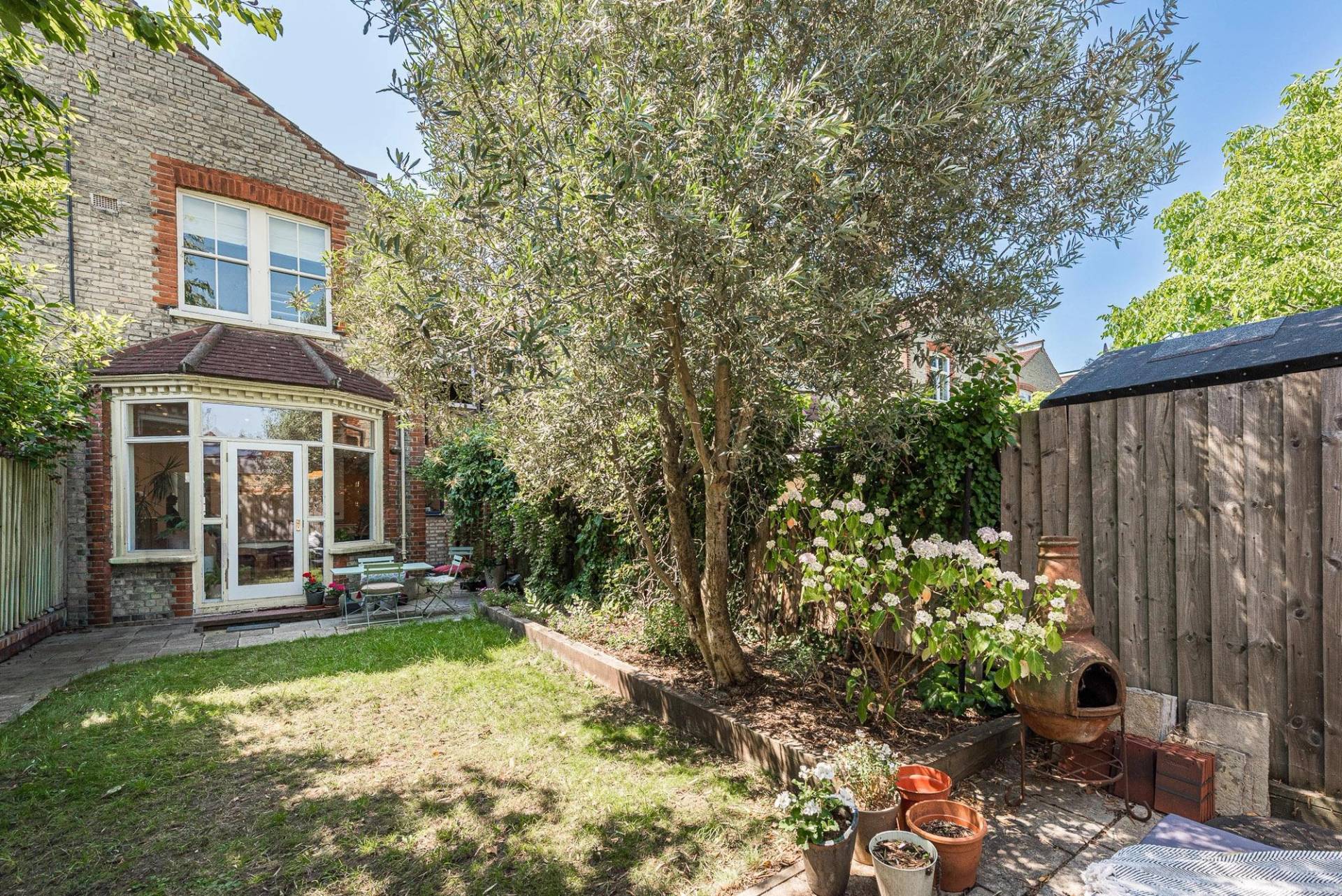 Secluded sunlit rear garden location with a lush green lawn and mature olive trees in Barnet, N11 via Love Locations.