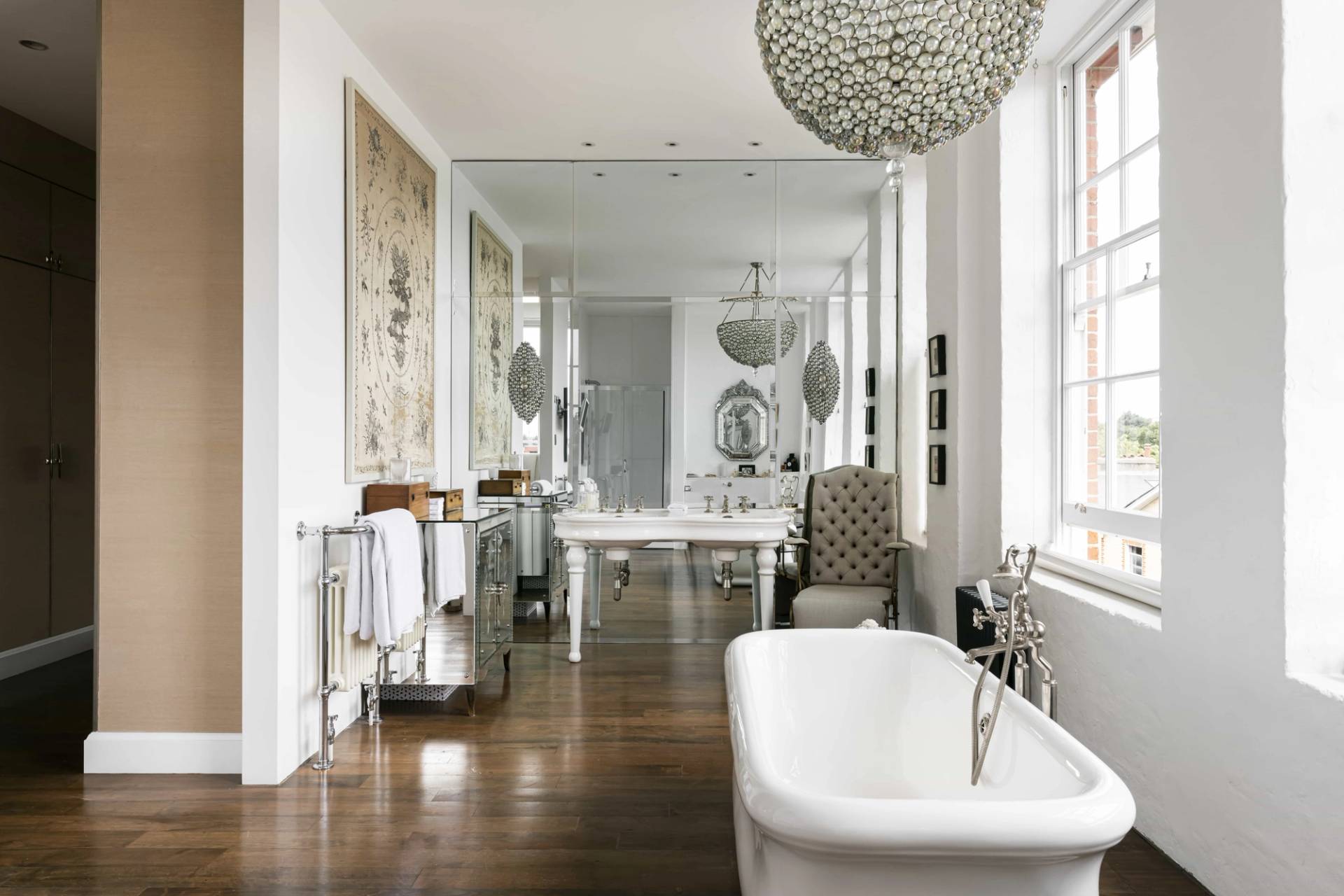 Grand bathroom featuring a white roll-top bath, mirrored wall panels, spherical crystal chandelier, and dark wood floors, Love Locations SW11.