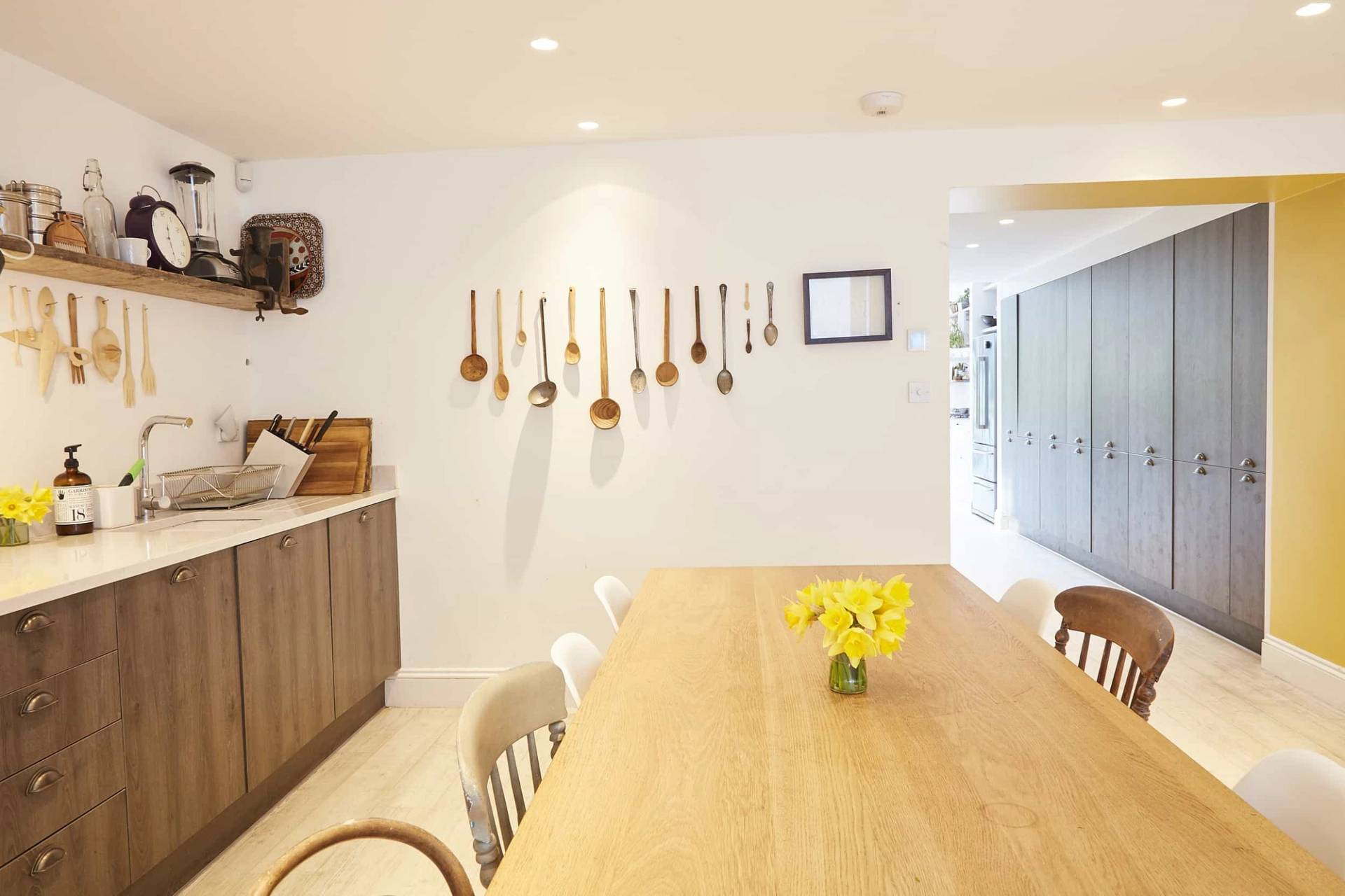 Close-up perspective of a solid oak dining table styled with yellow flowers and a hanging wooden spoon wall collection, Love Locations N5.