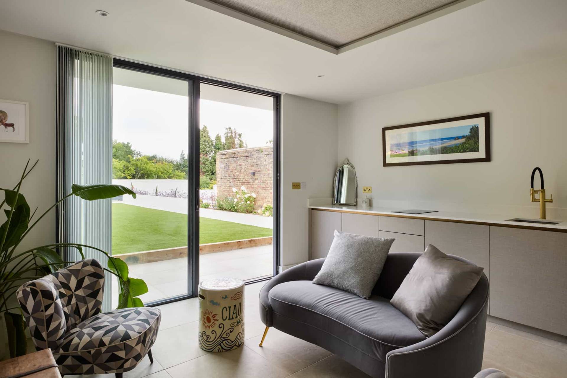 Cozy TV lounge or snug featuring a low-profile grey sofa, floor-to-ceiling glass doors, and garden views, ideal for domestic brand commercials, Love Locations KT22.