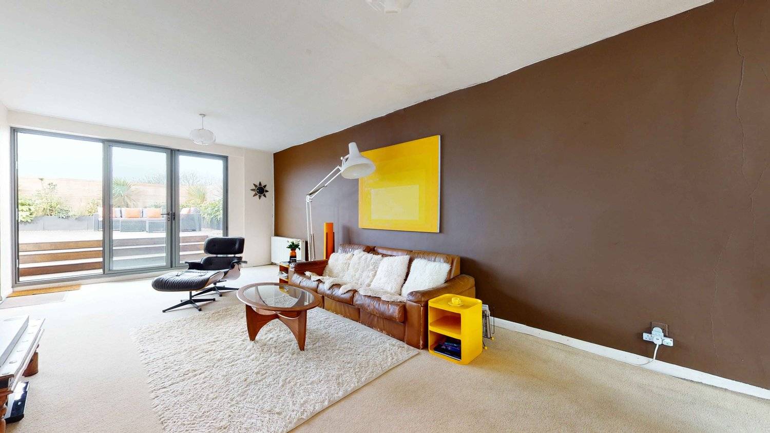 Spacious 1970s reception room with a large tan leather sofa, shag area rug, and yellow accent decor, Love Locations SW16.