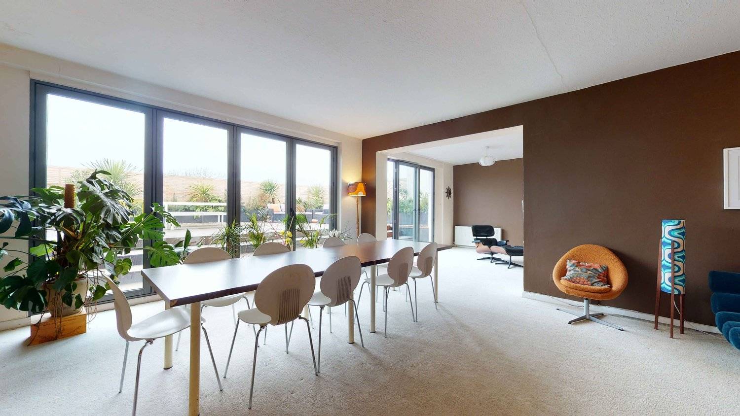 Mid-century modern dining room featuring a long conference table, white bucket chairs, brown wall panelling, and carpet flooring, Love Locations SW16.