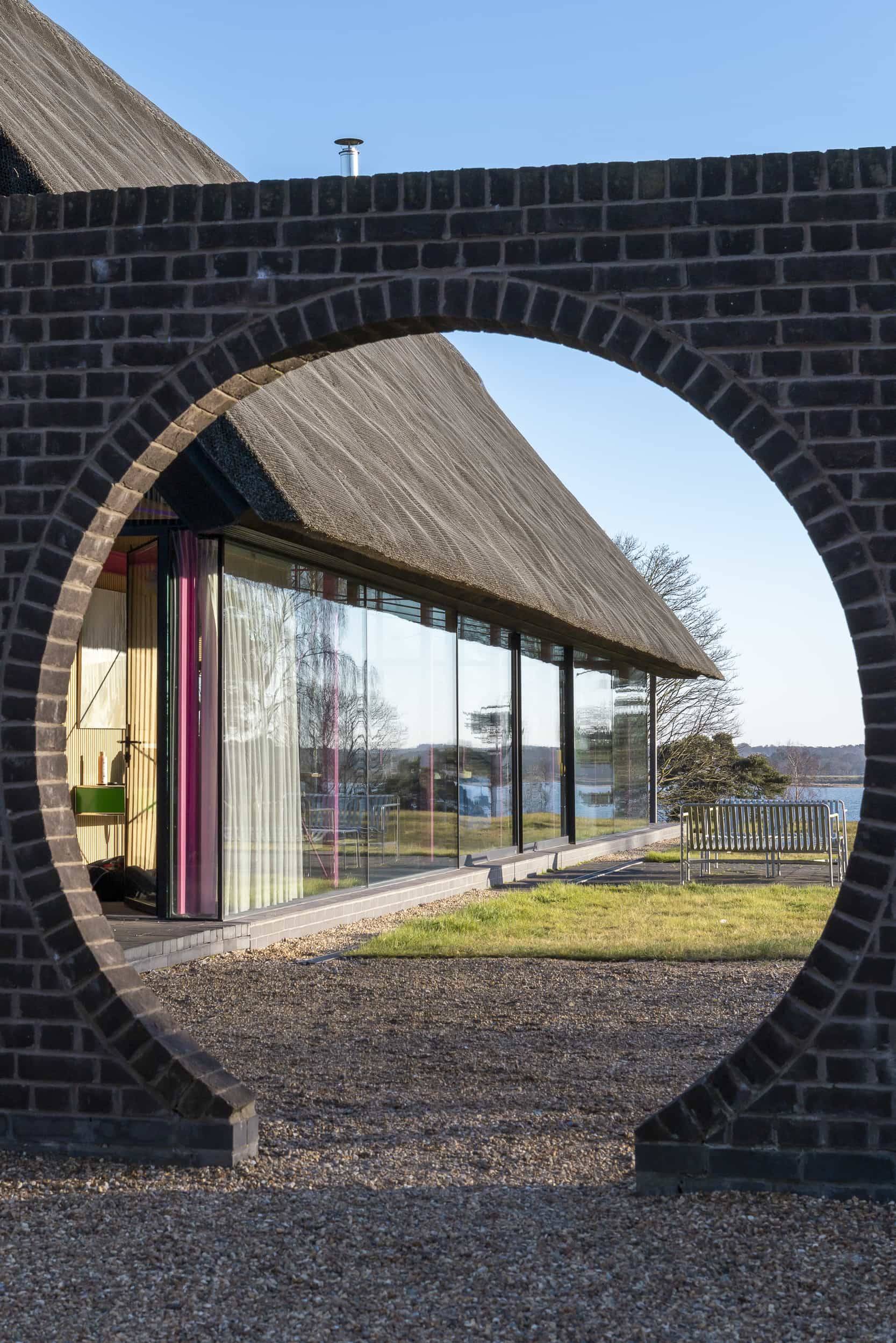 Circular brick wall archway framing a view of a modern glass longhouse with thatched roof in Saxmundham, Love Locations IP17.
