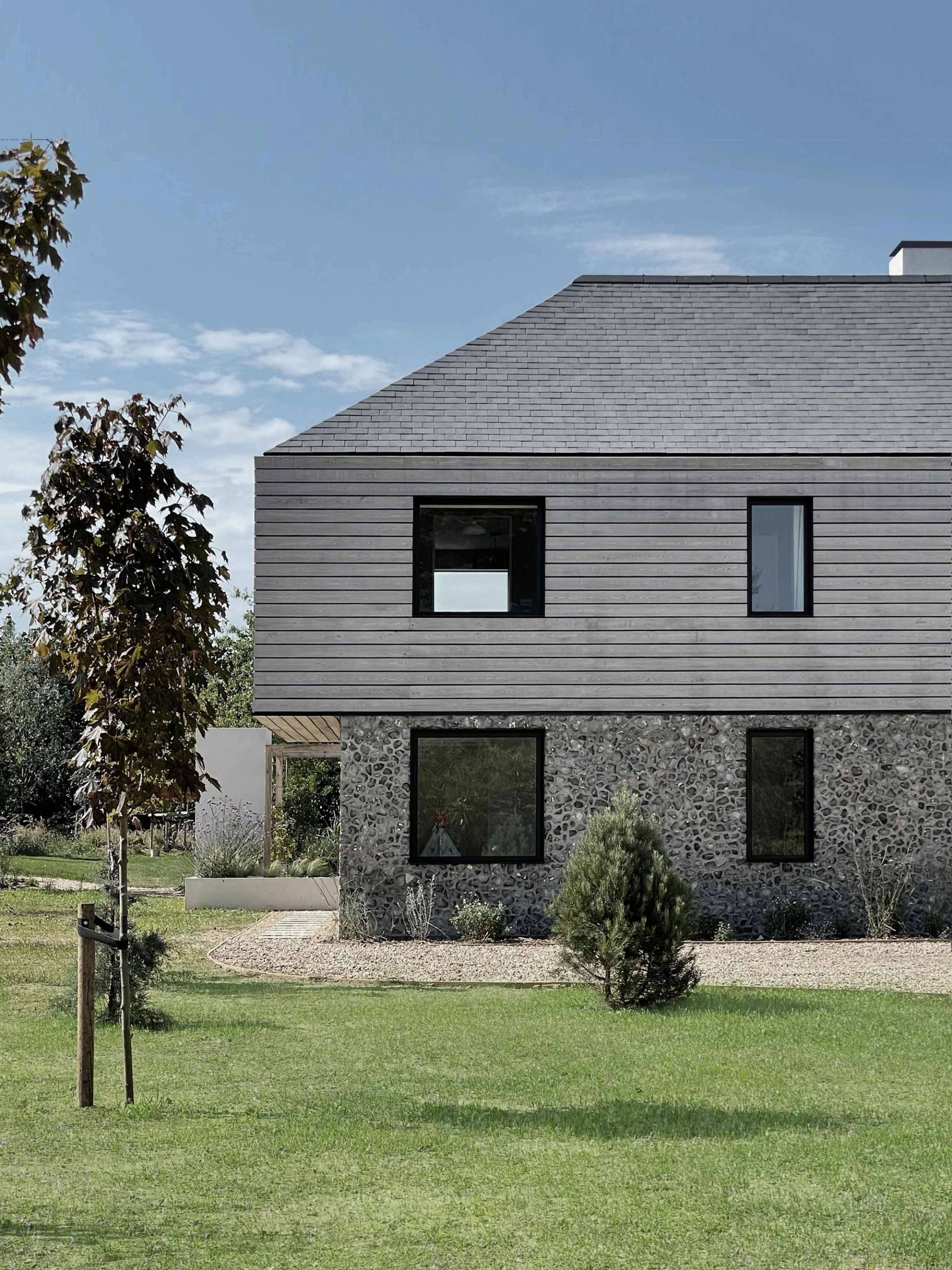 Exterior facade shot of a modern Chichester country home combining grey timber cladding, flint stone walls, and manicured lawns, ideal for TV drama establishing shots via Love Locations PO18.