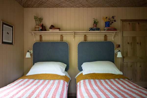 Twin bedroom setup inside a luxury timber cabin showing blue headboards, striped linens, and an overhead shelf, Love Locations.