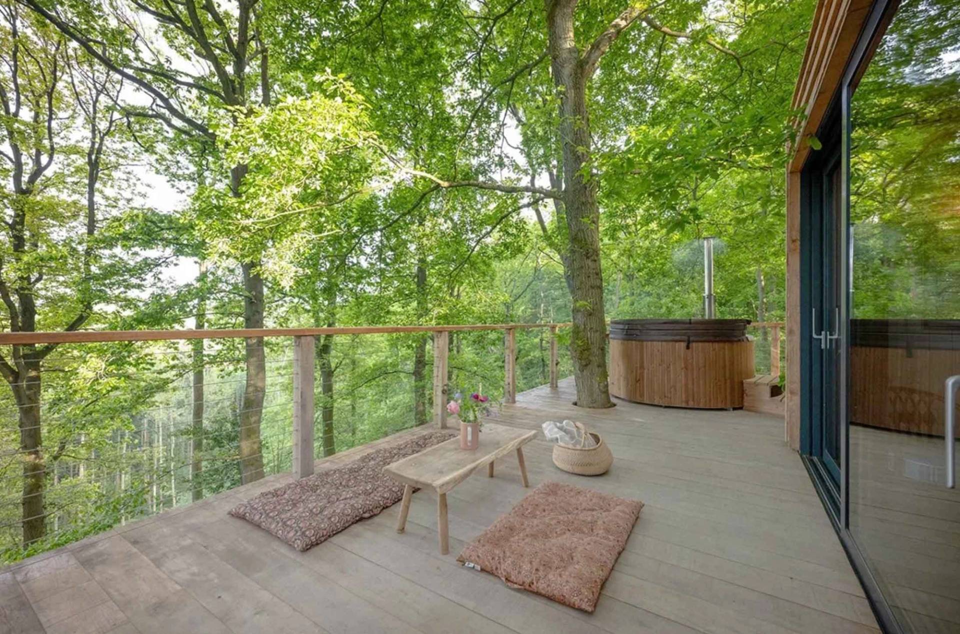 Outdoor wooden treehouse balcony deck equipped with floor cushions, a low coffee table, and an integrated hot tub, ideal for luxury glamping campaigns, Love Locations.