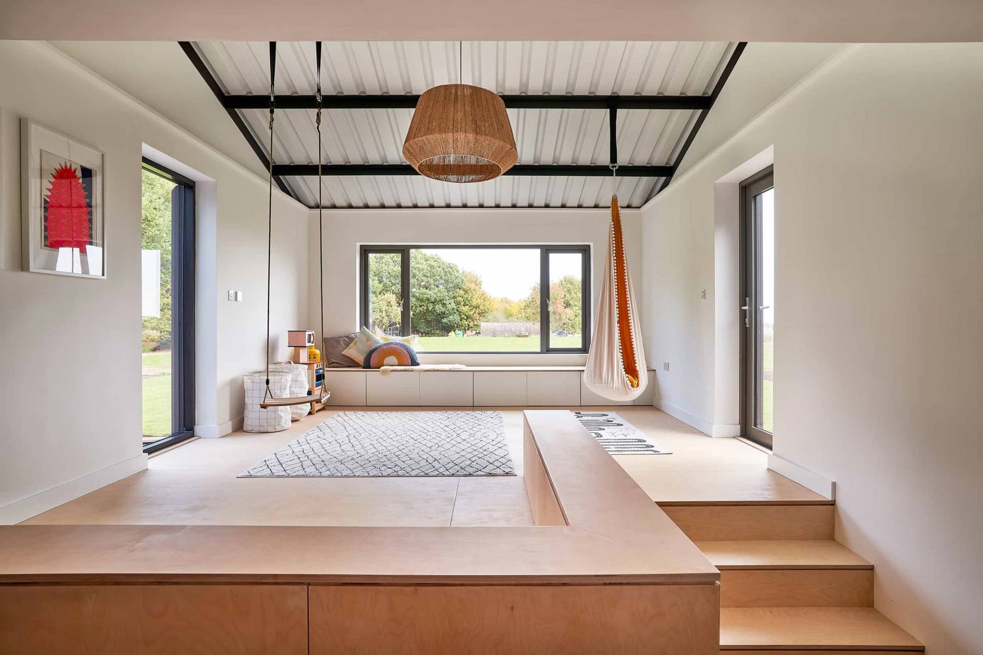 Sunken wooden lounge seating platform set under industrial black roof trusses with wide countryside window views, Love Locations IP30.