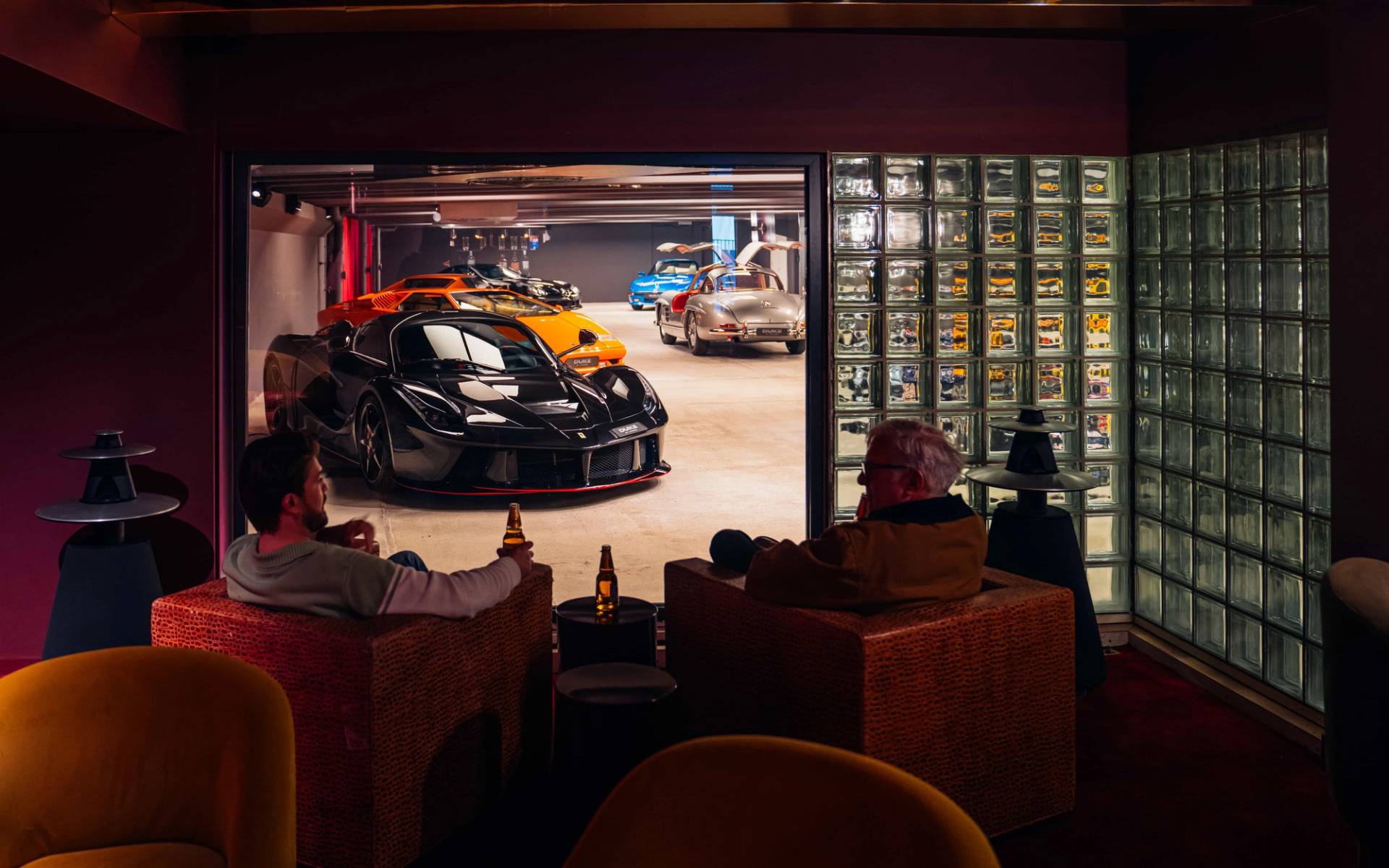Moody speakeasy bar lounge in Brentford TW8 from Love Locations, featuring warm leather armchairs looking through a window into a illuminated hypercar garage.