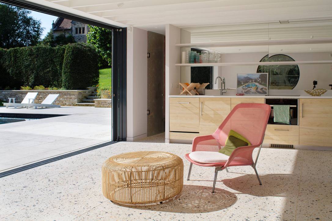 Pool house living space with pink armchair, woven pouf, terrazzo flooring, and sliding glass doors, Love Locations OX7.