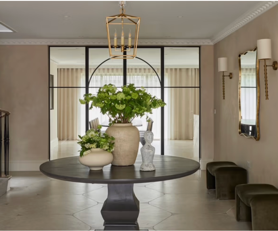 An elegant entrance hall with a round pedestal table styled with fresh flowers and decorative objects, black-framed glass doors, brass lighting, olive velvet benches, and soft neutral interiors creating a refined contemporary look.