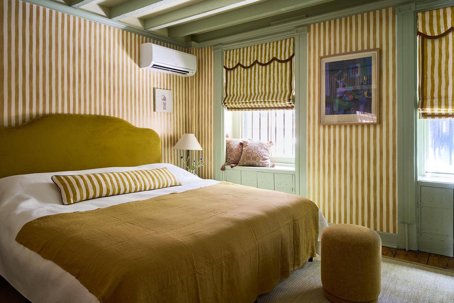 Warm yellow stripe wallpapered bedroom photoshoot location in Manhattan NY 10014 from Love Locations, featuring a padded mustard headboard, ochre duvet, and painted timber ceiling joists.