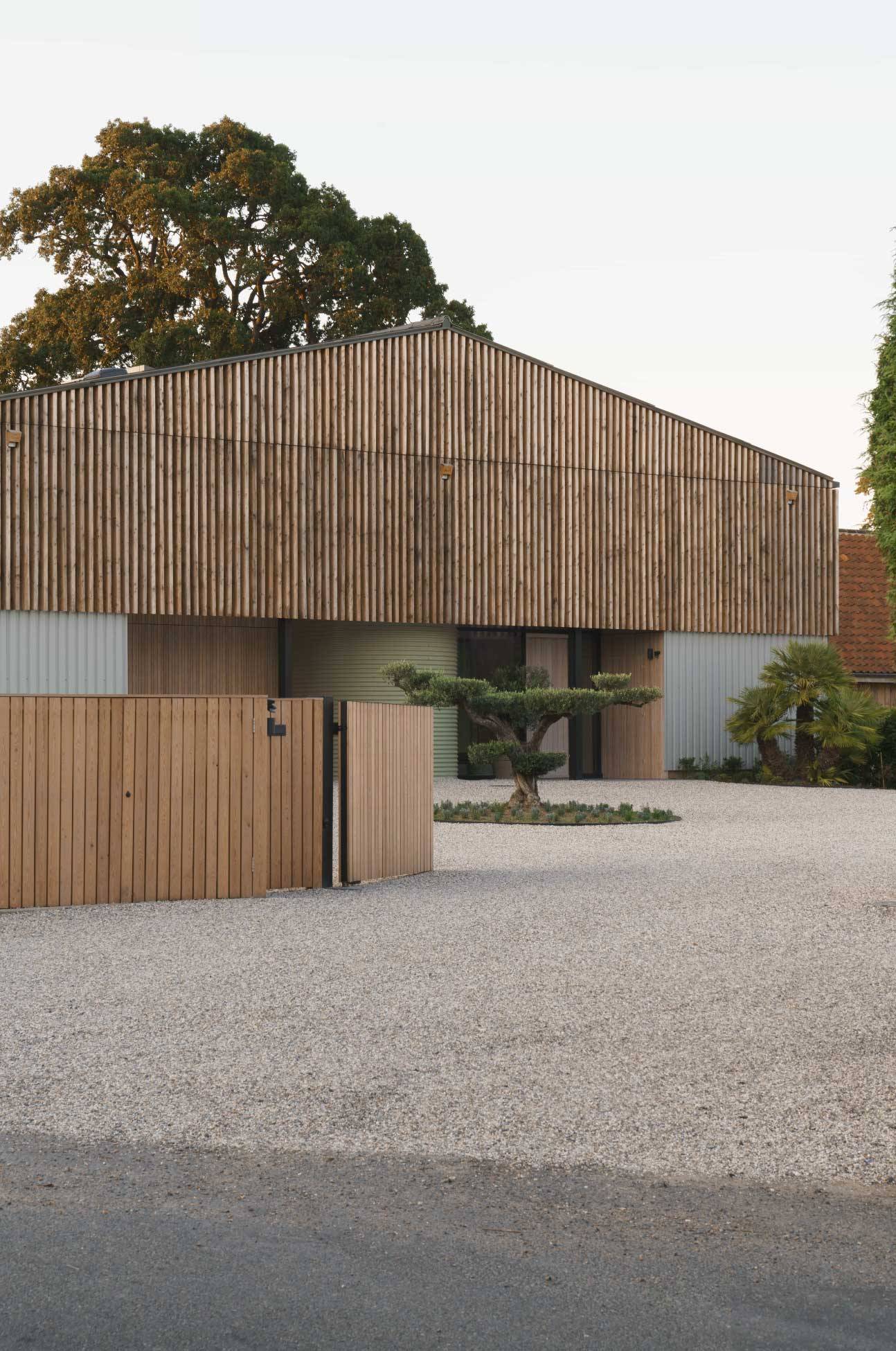 Gravel driveway courtyard featuring a timber-slatted architectural facade, timber fencing, and manicured trees, Love Locations NR9.