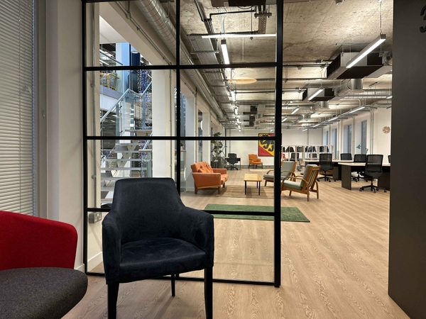 Open black crittall door framing a view into a bright, modern office space, with a red statement chair in the foreground.