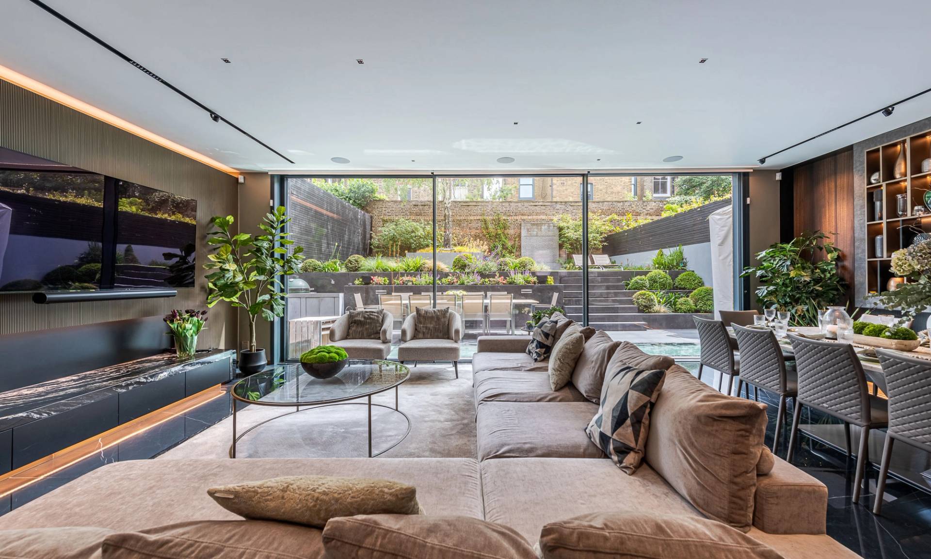 Low-profile grey sectional sofa set in a sunlit reception room overlooking a tiered garden terrace, Love Locations SW11.