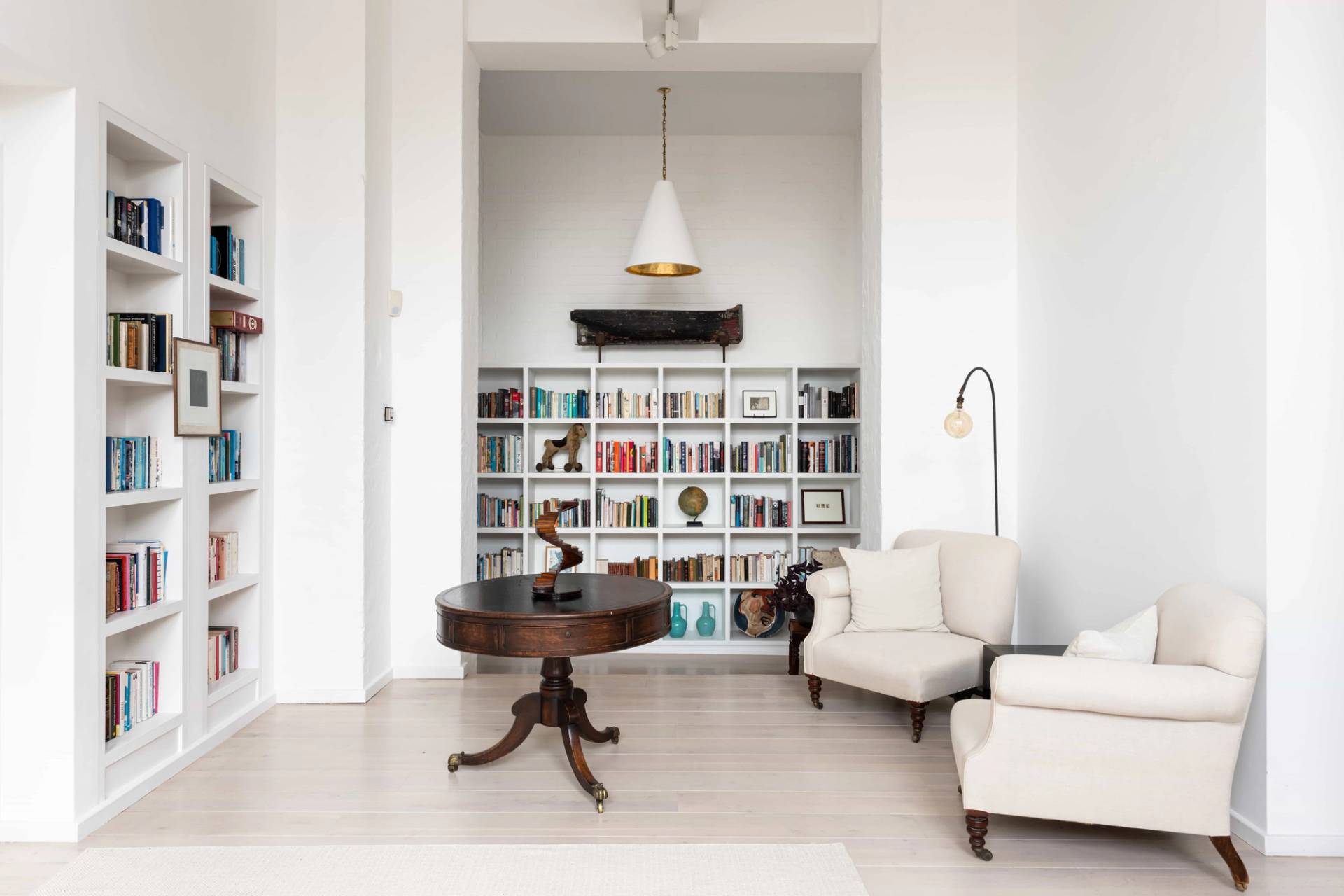 Reading corner setup featuring a dark wood round table, white armchairs, and floor-to-ceiling bookshelves in an arched alcove, Love Locations SW11.