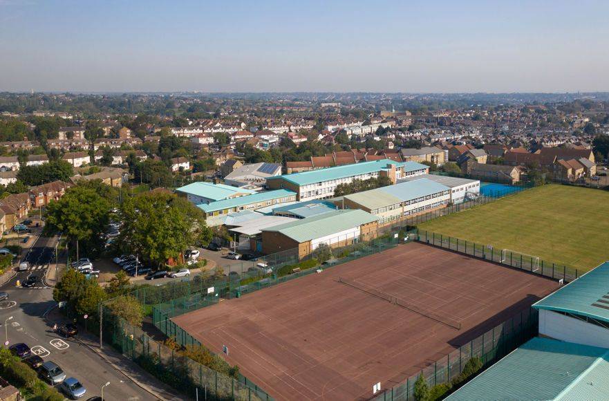 High-angle aerial drone view of the school campus buildings, outdoor sports courts, and surrounding North Finchley neighborhood through Love Locations.